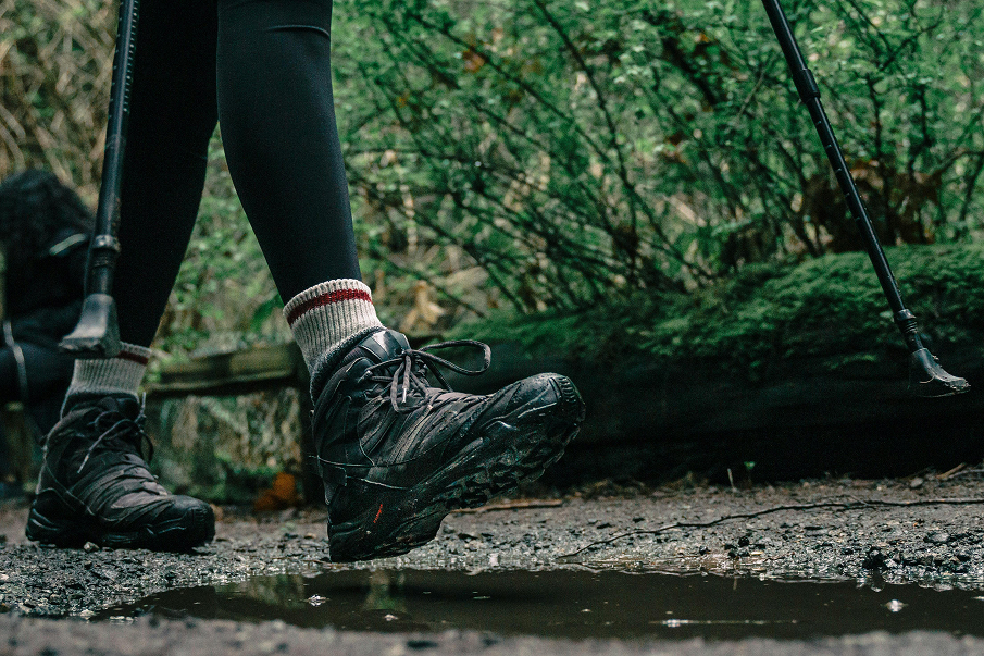 Zbliżenie na nogi osoby w butach trekkingowych idącej po leśnej ścieżce, z widoczną kałużą i kijkami trekkingowymi